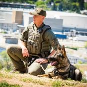 Un policier de la <abbr>GRC</abbr> en uniforme tactique est accroupi aux côtés d'un berger allemand, au sommet d'une colline.