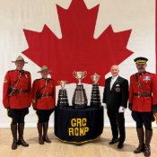Three RCMP officers and the Keeper of the Grey Cup stand with the Grey Cup, the Connaught Cup and the Minto Cup in front of a large Canadian flag.