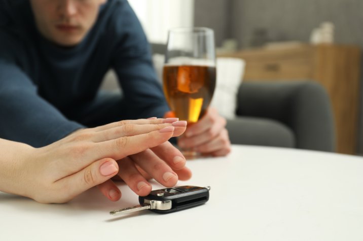 Close up of a set of car keys on a table in the foreground, with a person holding a glass containing an alcoholic beverage seated in the background.
