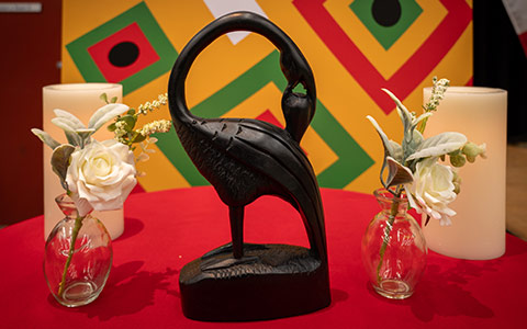 Un oiseau Sankofa, entouré de roses blanches et de bougies, est posé sur une table recouverte d'une nappe rouge.