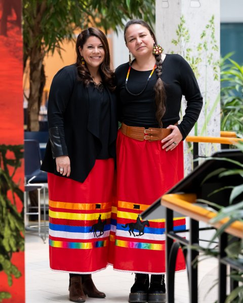 Deux femmes vêtues d'un chandail noir et d'une jupe longue rouge au motif de l'emblématique gendarme à cheval de la GRC et de rubans multicolores sourient.