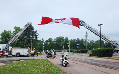 Motorcycles sit underneath a Canadian flag that’s being hoisted by firetrucks.