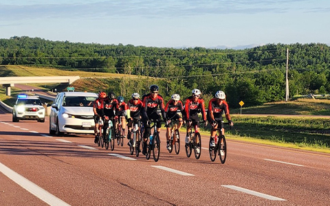 Several bicyclists are seen travelling along the road; an RCMP vehicle follows behind.