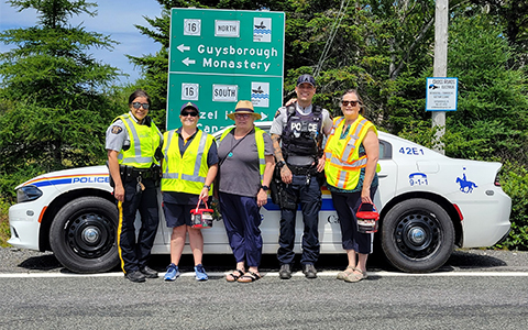 Five people including a couple of police officers, stand in front of a police car and highway signage.