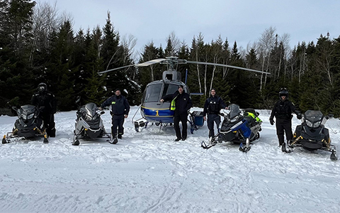 RCMP officers and snowmobilers stand alongside snowmobiles and a helicopter.