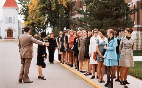 Des femmes sont alignées le long d'un trottoir. Derrière elles, un bâtiment en briques et une chapelle.