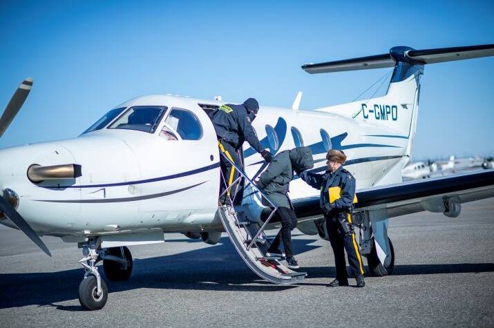 Two male RCMP officers escort a handcuffed individual with hood over his face away from an airplane.