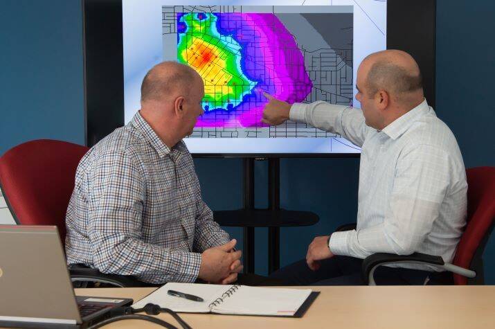 Two men sit while one points to a multi-coloured heat map on a screen.