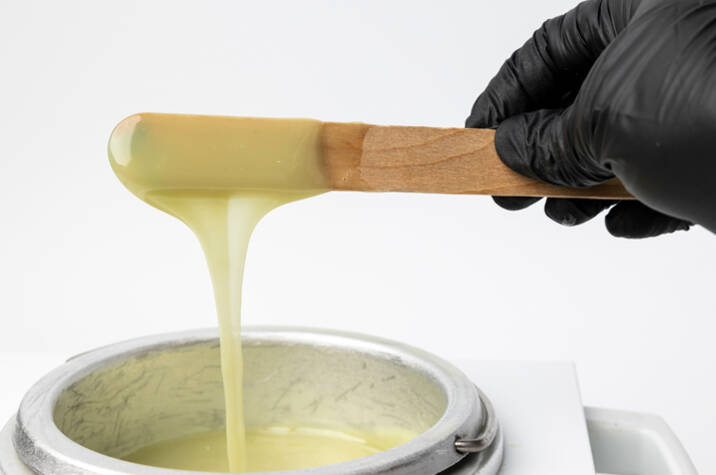 A black-gloved hand holds a stick dripping with light-yellow body wax.
