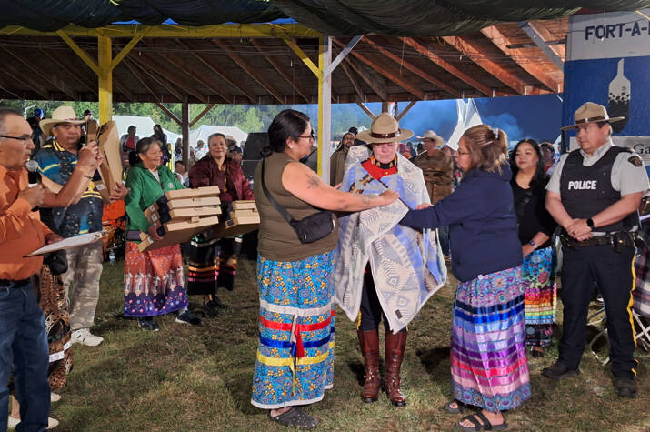 A man speaks to a crowd as an RCMP officer in red serge is wrapped in a star blanket by two women wearing Indigenous cultural ribbon skirts.