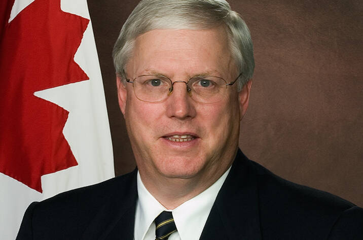 Portrait of a man wearing a dark suit and tie, with a Canadian flag behind him.