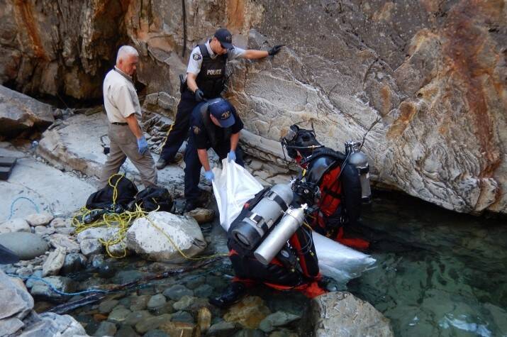 Two RCMP divers pull a bagged drowning victim from the water while male officers retrieve it from a rocky shore.