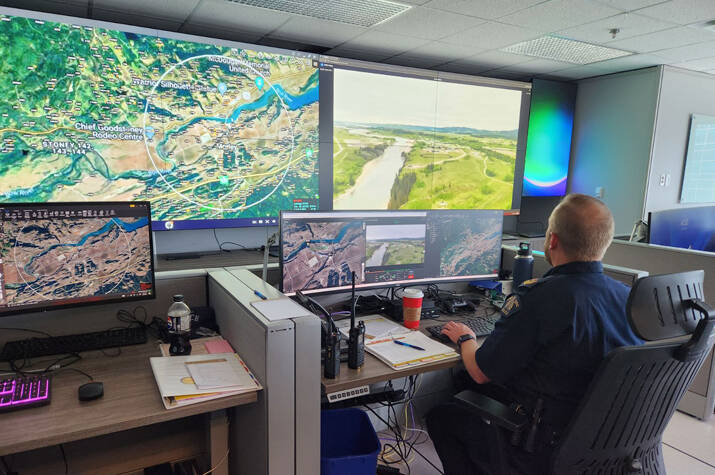 A uniformed RCMP officer sits in a chair inside a room filled with television screens and computer monitors. On the screens are a map and an aerial view of a landscape.