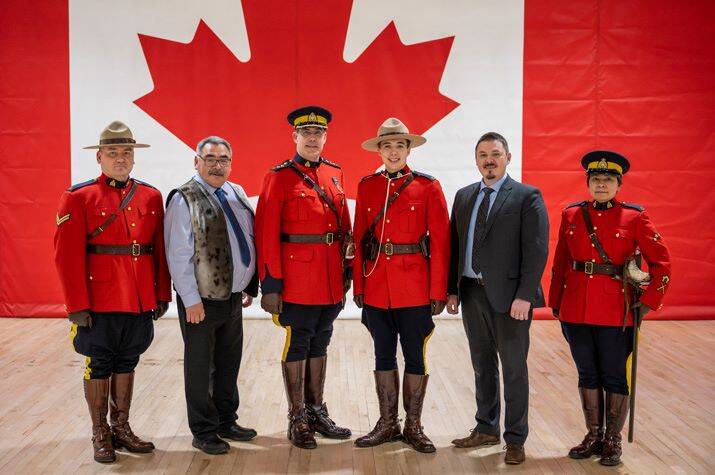 Four male and female officers wearing Red Serge pose in front of a large Canadian flag wall mural. Two men in business attire, stand in the row with them.