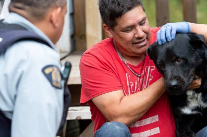 Police officer speaks with seated man embracing a big black dog.