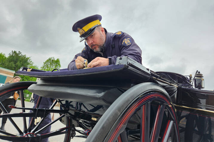 A man in blue coveralls leans over the back of a horse carriage polishing the black exterior.