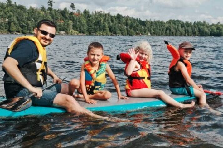 Three children and a man in floatation devices sit on top of a paddle board