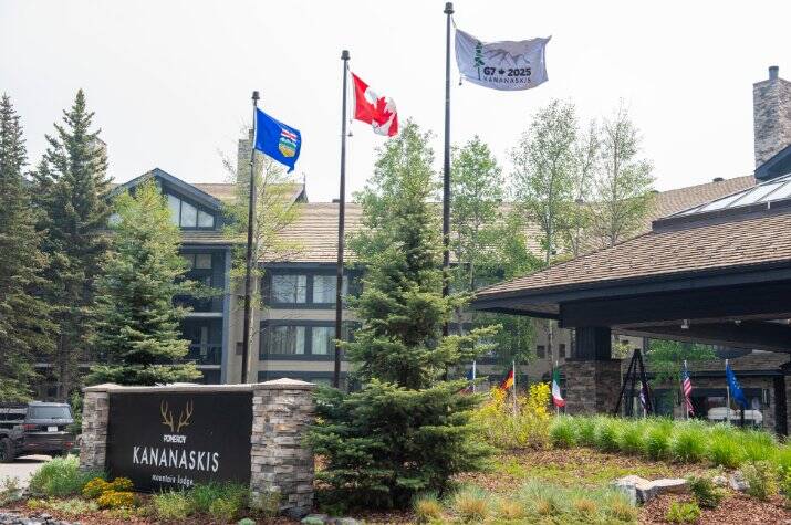 Building surrounded by evergreen trees. Three large flagpoles at the entrance display the Canadian, Albertan and G7 2025 flags, with smaller G7 country flags in the distance.