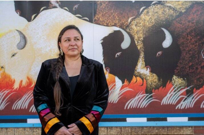 A woman with black hair and along braid stands in front of a mural with bison.